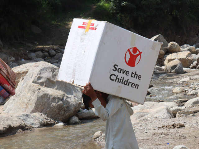 Two men carry supplies across a stream. One carries textiles, another has a box that reads "Save the Children"